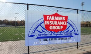 Blue gradient mesh banner hanging on a chain-link fence. Red and white logo in the middle is "FARMERS INSURANCE GROUP". 