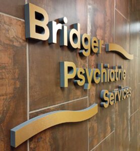 Light brown lettering "Bridger Psychiatric Services" positioned on a darker wood paneled wall.